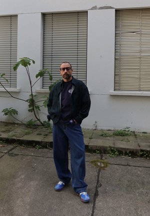 Hombre con gafas que lleva chaqueta oscura, camisa negra, vaqueros anchos azules y zapatillas azules, de pie al aire libre junto a un edificio blanco con contraventanas cerradas.