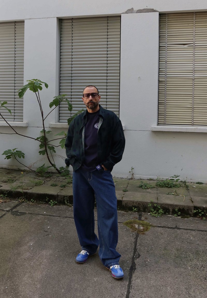 Homme avec des lunettes portant une veste sombre, une chemise noire, un jean large bleu et des baskets bleues, debout à l'extérieur près d'un bâtiment blanc avec des volets fermés.