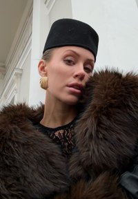 Brun pelsfrakk med svart blonder topp og svart filthatt. Har gullringøreringer og en teksturert pelsdetalj på frakken.