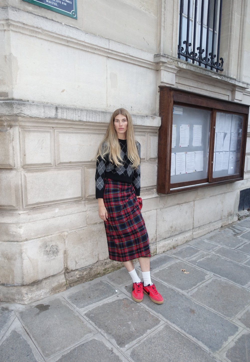 Black and grey argyle sweater paired with a red plaid wrap skirt. The model wears red trainers and white socks, standing against a stone wall.