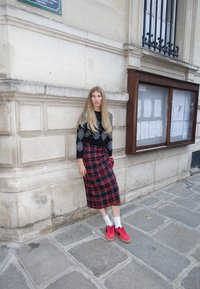 Black and grey argyle sweater paired with a red plaid wrap skirt. The model wears red trainers and white socks, standing against a stone wall.