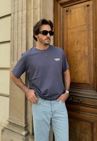 Σκούρο μπλε βαμβακερό t-shirt με λευκό λογότυπο στην αριστερή πλευρά του στήθους, συνδυασμένο με ανοιχτόχρωμα τζιν. Το φόντο απεικονίζει μια ξύλινη πόρτα.