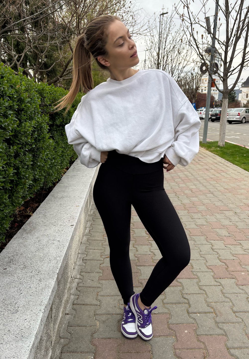 Loose-fitting white sweatshirt, high-waisted black leggings, and purple trainers with white details. Outdoor setting featuring pavement and greenery.