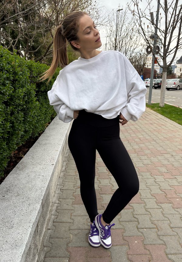 Sudadera blanca suelta, leggings negros de cintura alta y zapatillas moradas con acentos blancos. Entorno exterior con pavimento y zonas verdes.