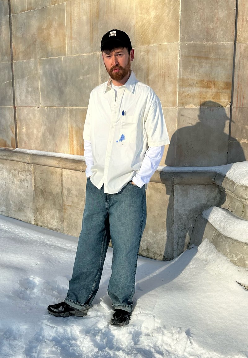 Mann steht im Schnee, trägt ein weißes Langarmshirt unter einem kurzärmligen Shirt, blaue Jeans, eine schwarze Kappe und schwarze Schuhe, neben einer Steinmauer.