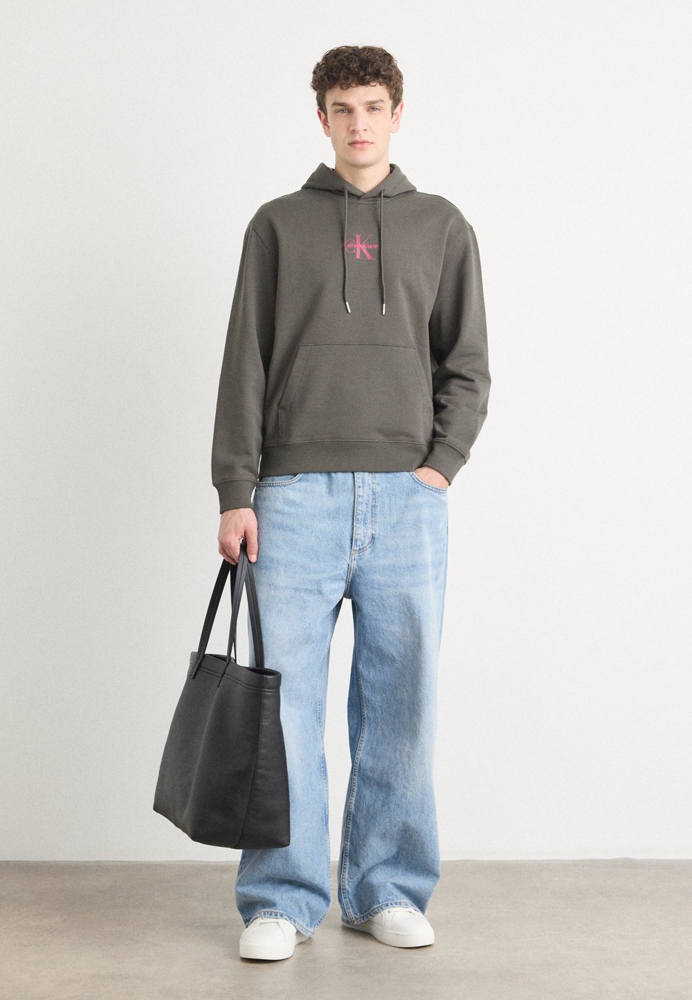 Pilkas džemperis su logo, priekinė kišenė ir raišteliai; laisvi šviesiai mėlyni džinsai; juoda rankinė; balti sportbačiai. Paprastas, kasdieniškas apranga.