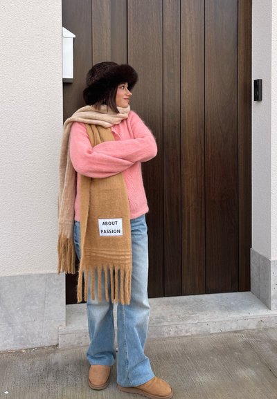 Suéter rosa suave, bufanda beige con flecos, jeans azules y botas marrones. El fondo presenta una puerta de madera con una pared de color claro.