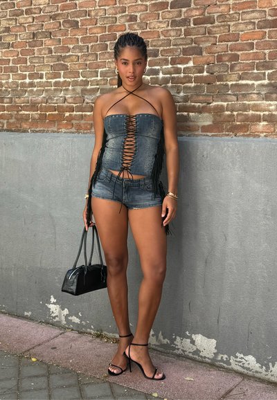 Top sin mangas de denim con detalle de cordones en el frente y flecos, combinado con pantalones cortos azul oscuro y sandalias negras con tiras. También incluye un pequeño bolso negro.