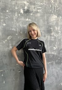 Svart kortärmad Adidas-t-shirt med gröna accenter och stjärndesign; bärs med en strukturerad svart kjol. Grå strukturerad bakgrund.