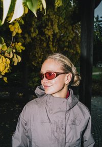 Grå vattentät jacka med hög krage, i kombination med röda solglasögon. Anmärkningsvärda detaljer inkluderar droppar på jackans yta och en strukturerad finish.