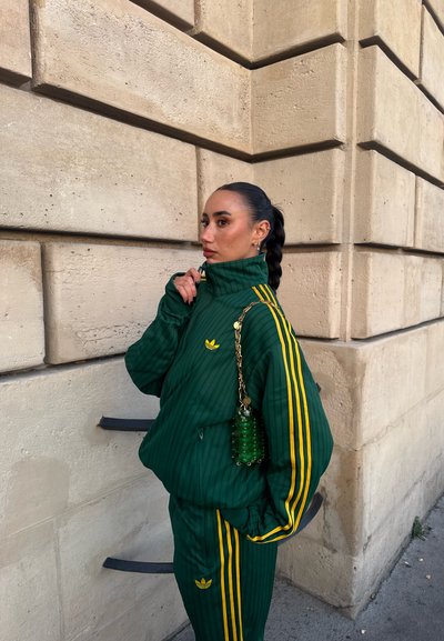 Mujer con el cabello trenzado que lleva un chándal verde de Adidas con rayas amarillas, parada frente a una pared de piedra beige sosteniendo un bolso verde con cadena dorada.