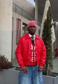 Casaco bomber vermelho com design em relevo, sobre um suéter padrão vermelho e branco, combinado com jeans de ganga azul e um gorro listrado.