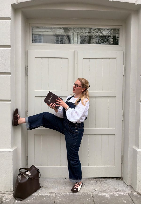 Kobieta w okularach i dżinsowym ogrodniczku opiera się o białe drzwi z jedną uniesioną nogą, czyta książkę, obok na ziemi leży brązowa torba.