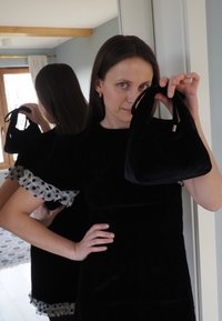 Sac à main en velours noir avec une forme sculptée et des poignées courtes. Fond avec un mur bleu clair et des rideaux blancs. Détails à pois sur les manches.