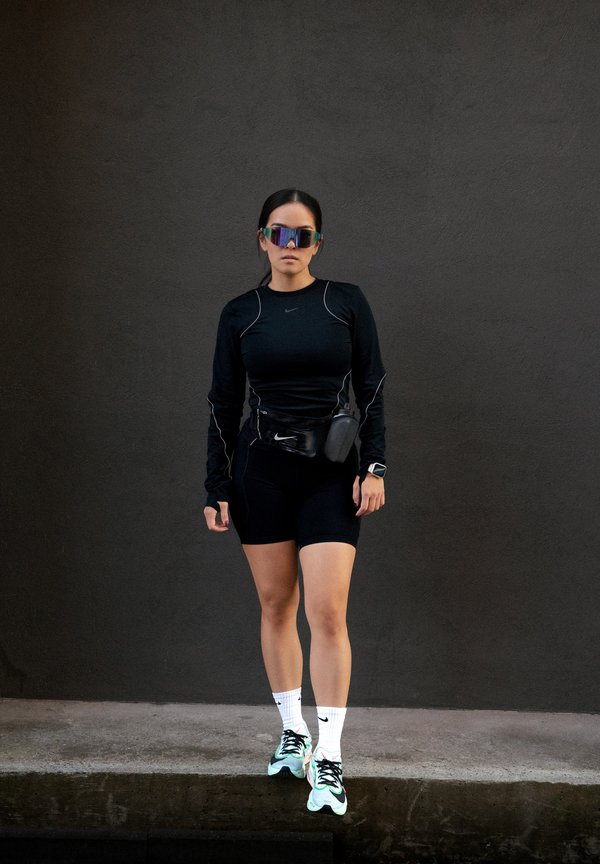 Mujer con conjunto deportivo negro, gafas de sol reflectantes, una riñonera y calcetines blancos de Nike con zapatillas verdes y negras, de pie frente a una pared oscura.