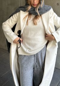 Manteau en laine blanche, pull beige et pantalon de survêtement gris. Coupe longue et structurée avec une texture dans les tissus et une écharpe grise portée sur l'épaule.