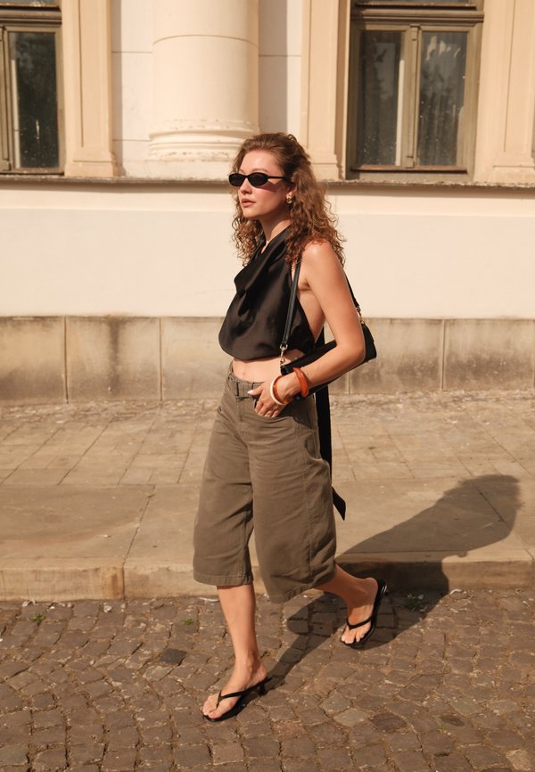 Frau mit lockigem Haar, die ein schwarzes ärmelloses Oberteil, Sonnenbrille, weite khakifarbene Shorts und schwarze Sandalen trägt, geht auf einer gepflasterten Straße.
