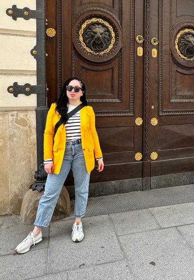 Blazer amarillo sobre una camisa a rayas, jeans azul claro y zapatillas blancas. El fondo presenta puertas de madera ornamentadas con medallones de león.