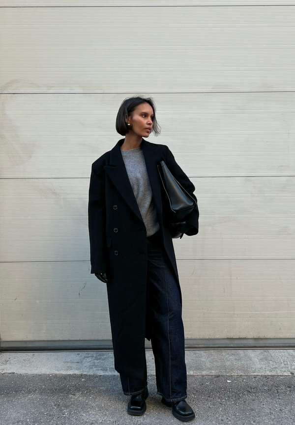 Mustat kaksiriviset takki harmaan neuleen päällä, yhdistetty tummiin farkkuihin ja mustiin saappaisiin. Käsissä tyylikäs musta käsilaukku.