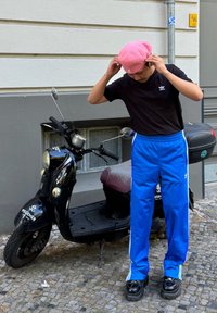 Melna Adidas T-kreklā, zili sporta bikses ar baltām sānu svītrām, rozā berete, stāv blakus melnai motociklam ar sarkanu sēdekli.