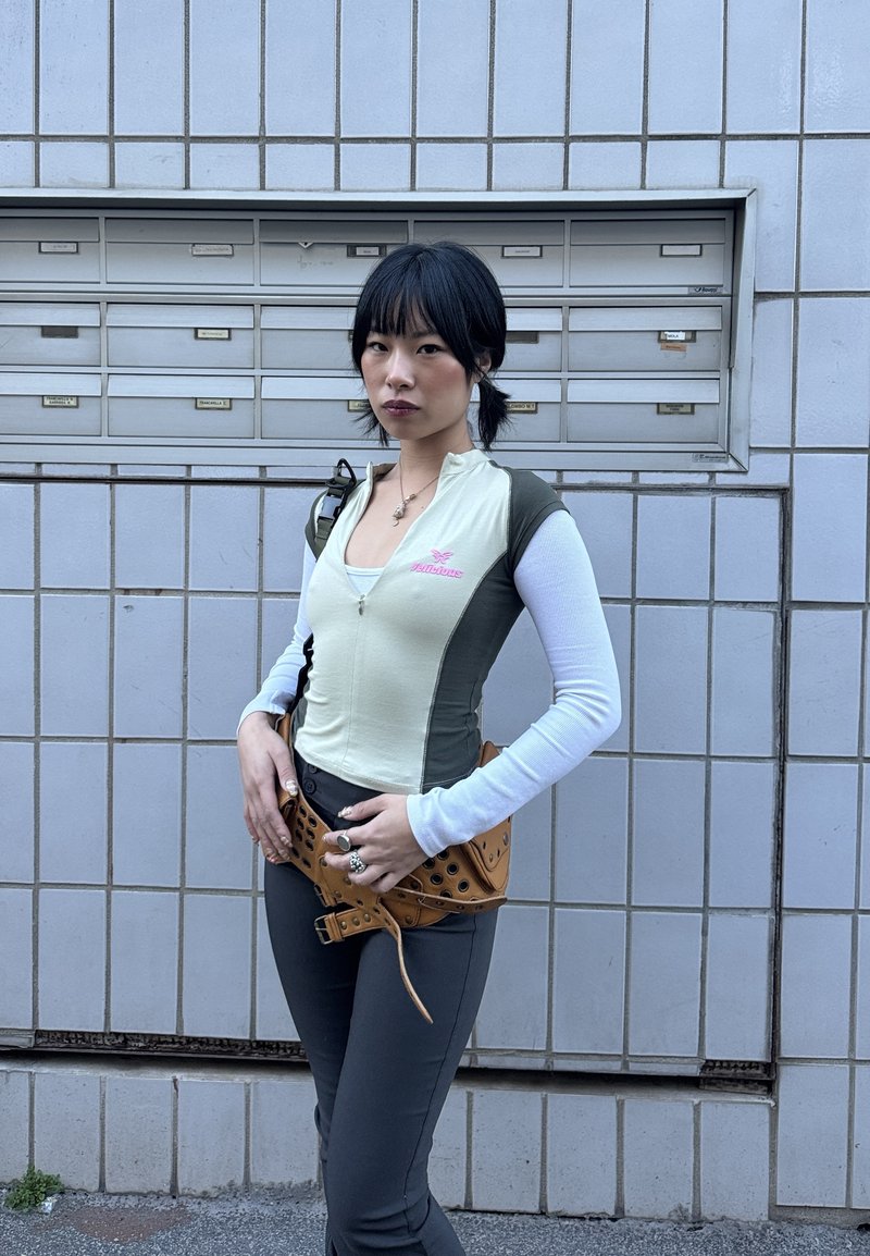 Jeune femme aux cheveux noirs coiffés en couettes, debout devant un mur carrelé avec des boîtes aux lettres, portant un haut ajusté à plusieurs couches, un pantalon foncé et une ceinture-sac marron.