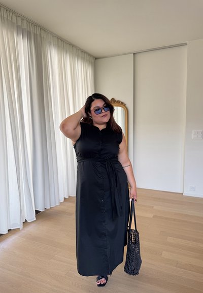 Vestido maxi negro sin mangas con botones y cintura anudada. Bolso negro texturizado y gafas de sol de gran tamaño. Suelo de madera clara.