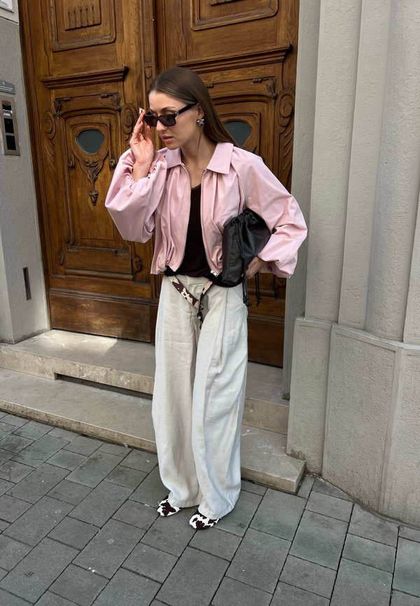 Femme ajustant ses lunettes de soleil, portant une veste rose, un haut noir, un pantalon blanc large, des chaussures à motif vache, tenant un sac noir près d'une porte en bois ornée.