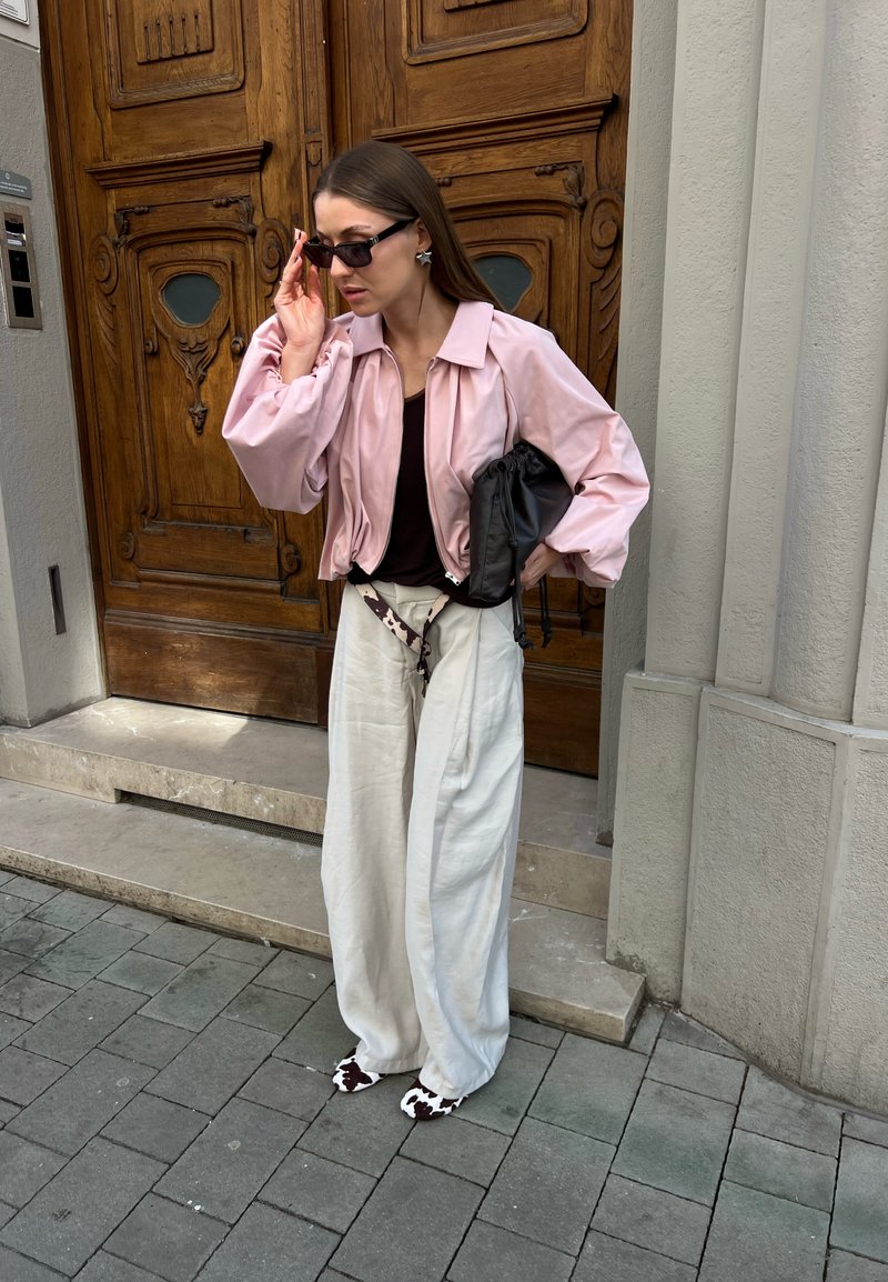 Femme ajustant ses lunettes de soleil, portant une veste rose, un haut noir, un pantalon blanc ample, des chaussures à motif vache, tenant un sac noir près d'une porte en bois ornée.