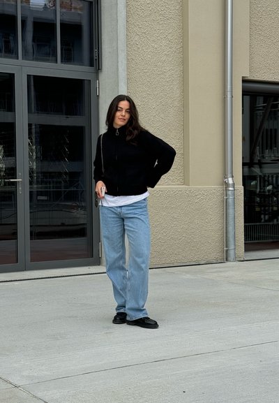 Sudadera negra con cremallera, camiseta blanca debajo, vaqueros de pierna ancha azul claro, zapatos negros. De pie cerca de grandes puertas de vidrio con una pared texturizada.