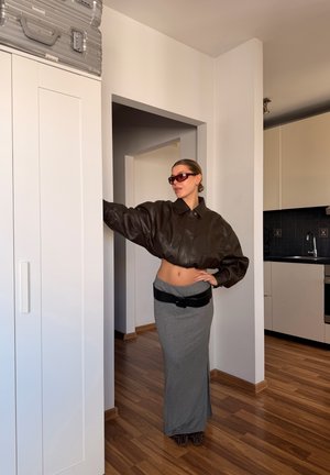 Mujer con gafas de sol, chaqueta marrón corta, falda gris y cinturón negro, de pie en el interior junto a un armario blanco en una habitación con suelo de madera.