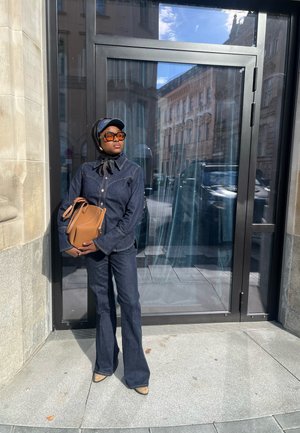 Jeansjacke und ausgestellte Jeans in dunklem Waschton, kombiniert mit einem schwarzen Schal und Sonnenbrille. Hält eine braune Lederhandtasche. Modernes urbanes Setting.