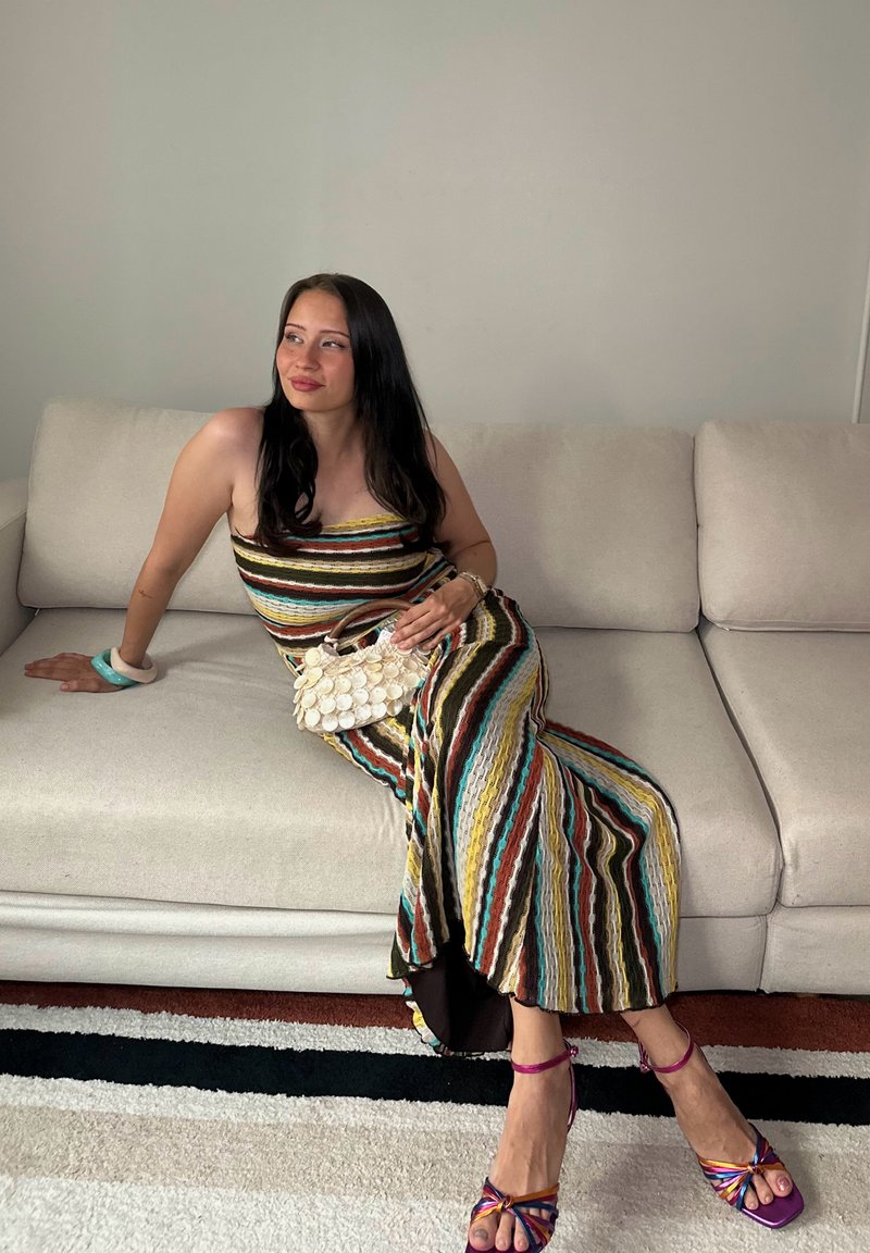 Femme portant une robe à rayures multicolores et des talons violets, assise sur un canapé beige, tenant un sac à main blanc en forme de coquillage, regardant sur le côté.