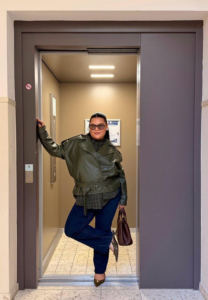 Femme portant des lunettes de soleil et une veste verte se tient décontractée dans l'encadrement d'un ascenseur éclairé beige, tenant un sac à main marron et touchant le cadre de la porte.