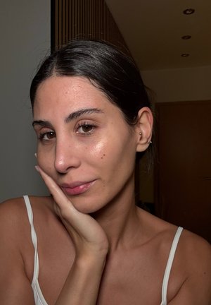 Portrait of a person with medium skin tone, dark hair, and a subtle sheen on the face. Wearing a white strap top, lightly touching their cheek.