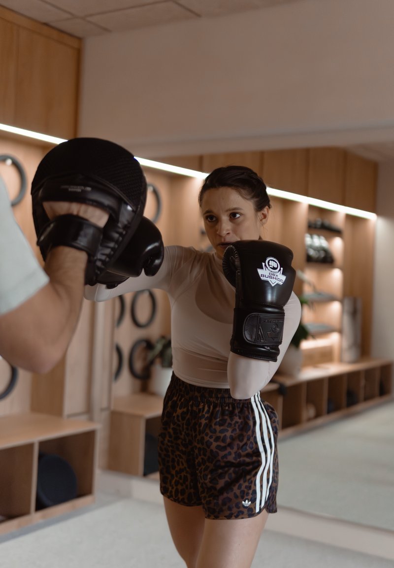 Femme portant des gants de boxe s’entraîne à donner des coups sur des mitaines tenues par un entraîneur dans une salle de sport moderne.