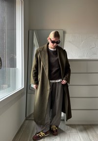 Olivgrüner Wollmantel über einem dunklen Pullover, heller Bluse, grauen Karohosen und braunen Sneakers mit gelben Schnürsenkeln. Spiegel und heller Hintergrund.