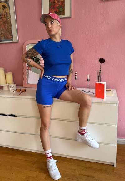 Top corto azul y shorts a juego con cinturilla "Nike Pro". Zapatillas blancas con acentos rojos. Fondo rosa con elementos decorativos.