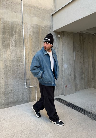 Chaqueta de mezclilla de tono azul claro, camiseta blanca, pantalones negros y gorro negro. Las zapatillas tienen un diseño en blanco y negro. Fondo de concreto.