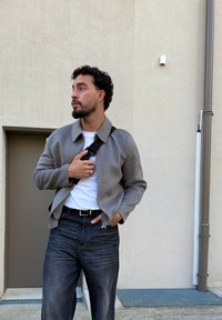 Göndör hajú férfi szürke zakót, fehér inget, sötét farmernadrágot és fekete keresztpántos táskát viselve áll egy bézs fal és zárt ajtó mellett.