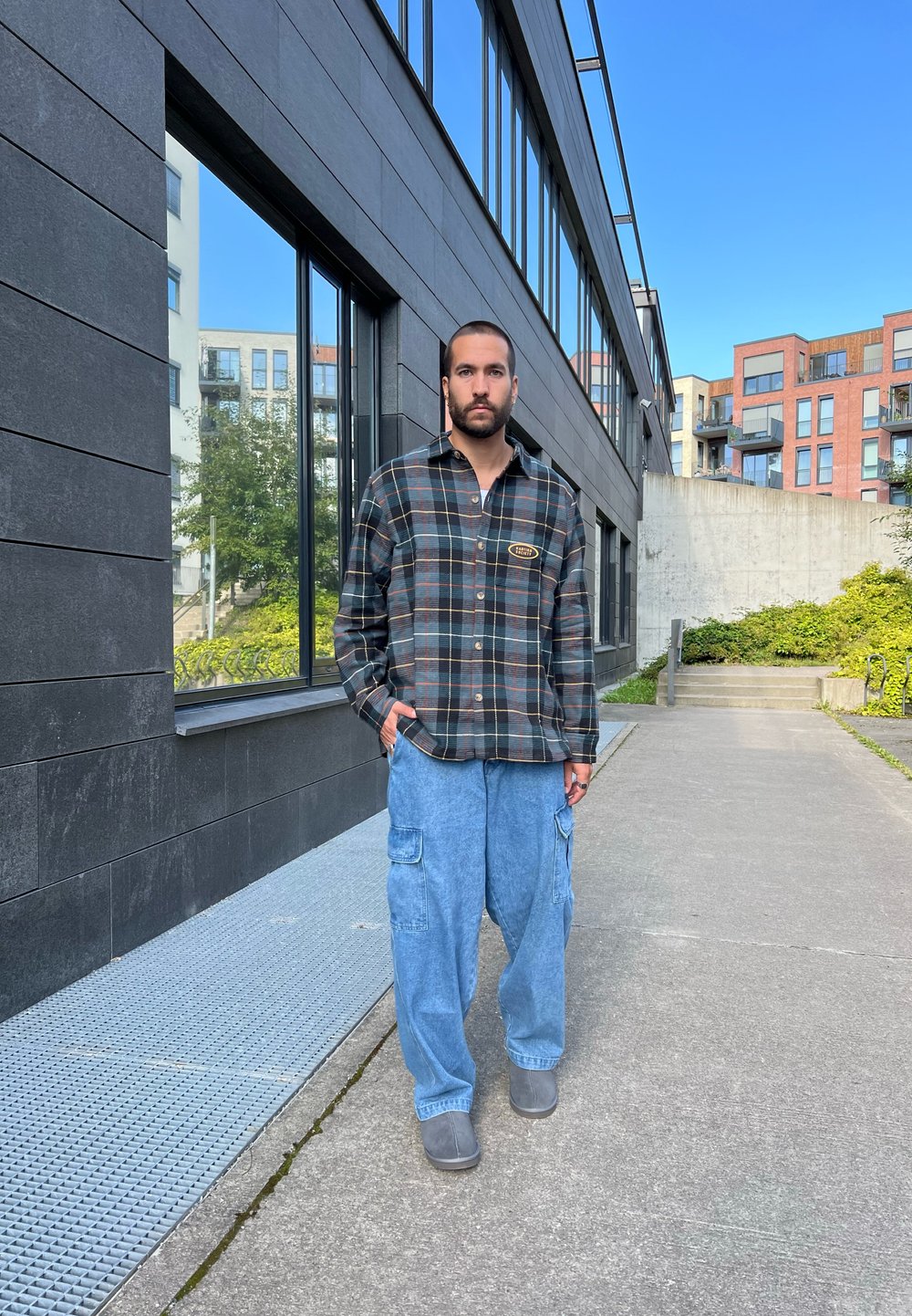 Camisa de flanela xadrez em cores escuras com um logótipo; calças cargo largas de tom azul claro; sapatilhas cinzentas sem atilhos; ambiente urbano exterior com edifícios modernos.