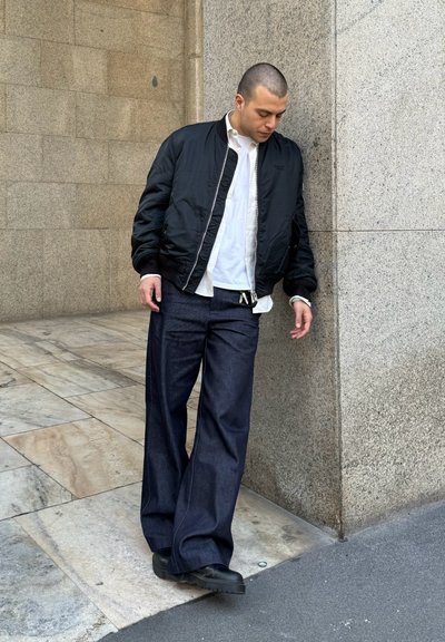 Chaqueta bomber negra sobre una camisa blanca, combinada con unos vaqueros de pierna ancha azul oscuro y zapatos negros. Texturas suaves y colores contrastantes.