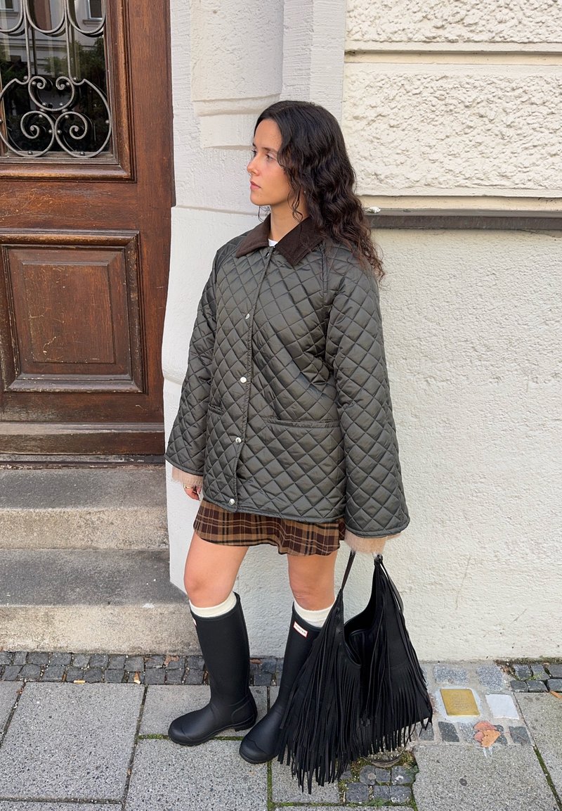 Quilted olive green jacket with brown collar, plaid skirt, black rubber boots, and a fringed black handbag, standing against a wooden door.
