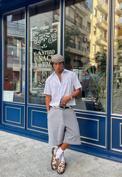 Hombre con una camisa blanca de manga corta, pantalones cortos a rayas y zapatos estampados, de pie junto a una tienda azul, con gafas y accesorios visibles.