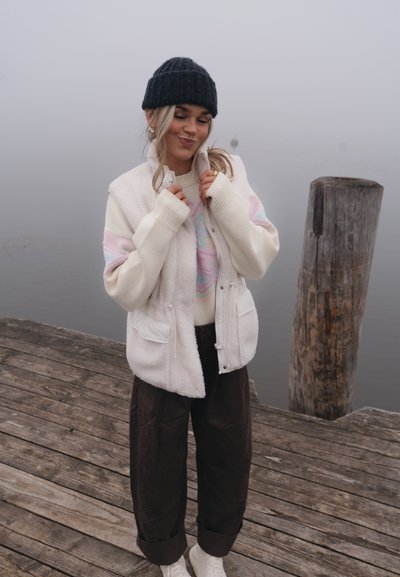 Chaleco blanco de forro polar con dos bolsillos delanteros, sobre un suéter con patrón en tonos pastel. Pantalones anchos marrón oscuro y un gorro negro de punto.