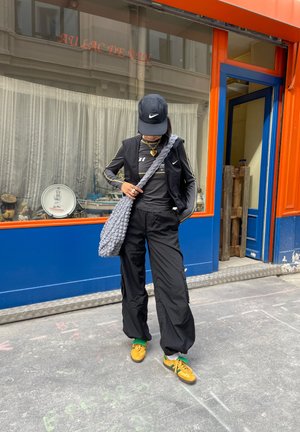 Chaleco atlético negro y pantalones, bolsa de punto gris y zapatos amarillos con cordones verdes. Gorra negra con un logo. Fondo neutro con acentos coloridos.