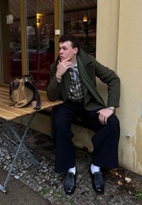 Ciemnozielony płaszcz, koszula w kratę, szary szalik, szerokie czarne spodnie oraz teksturowane czarne buty z białymi skarpetkami. Brązowa torba na drewnianym stole.