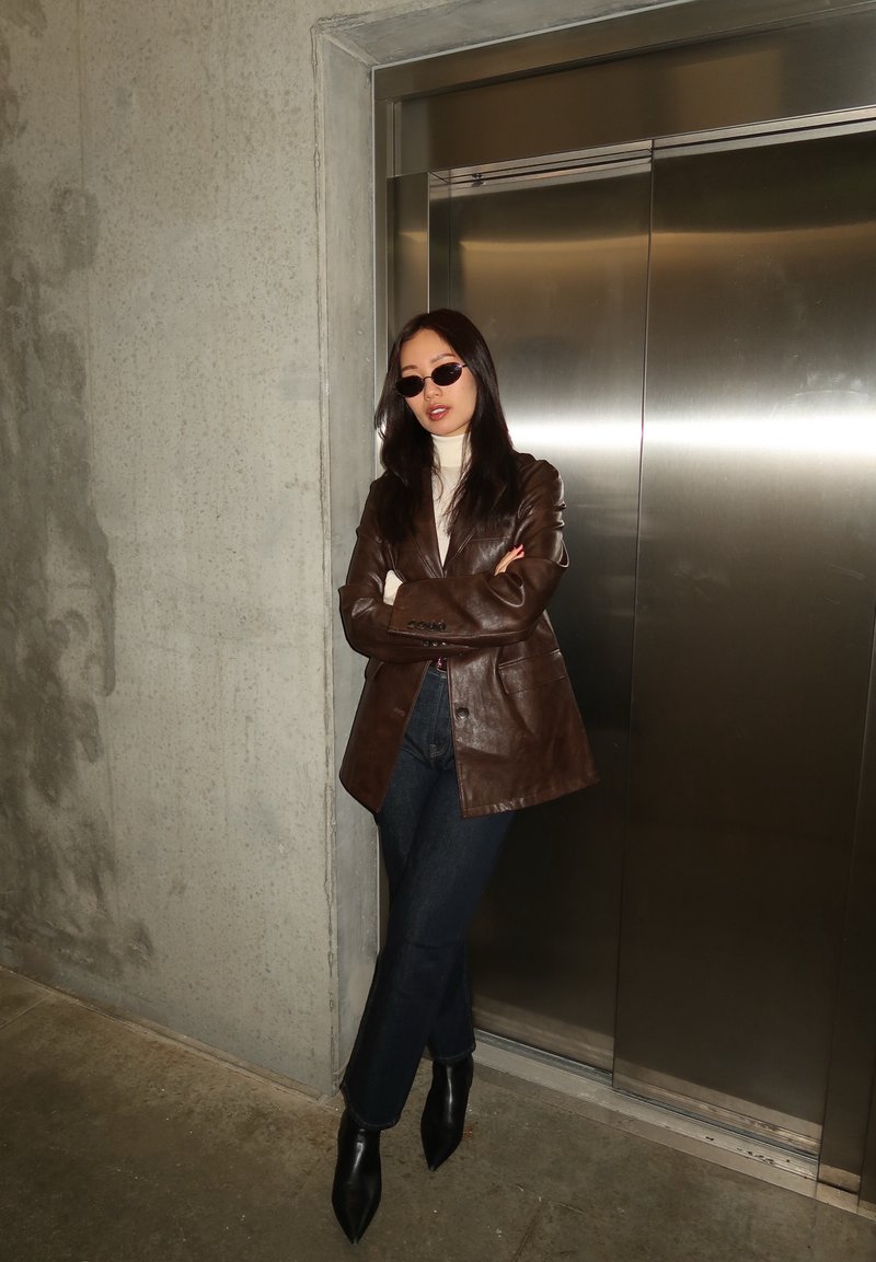 Femme portant des lunettes de soleil, un blazer en cuir marron, un col roulé blanc, un jean foncé et des bottes noires, debout les bras croisés devant des portes d'ascenseur fermées.