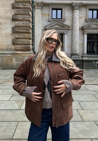 Jachetă din suede maro cu fermoar frontal, buzunare mari și guler gri, purtată peste un pulover cu model și asortată cu blugi negri.
