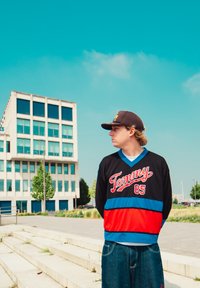 Černé tričko s červenými a modrými pruhy, s nápisem "Tommy 85". V kombinaci s modrými džínovými šortkami; vysoká budova v pozadí.