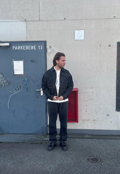 Chaqueta de mezclilla y pantalones en un lavado azul oscuro, combinados con una camisa blanca. De pie cerca de una pared y puerta grises con graffiti y un buzón rojo.